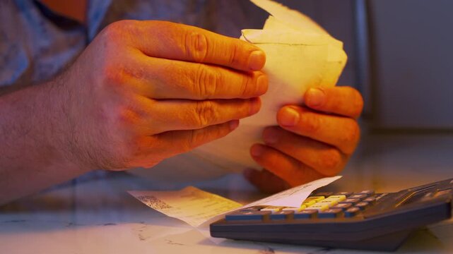 Low angle close-up of a man's hands sorting a stack of paper receipts under a desk lamp light. Cinematic close up shot of analyzing household expenses and choosing a receipt for financial audit.