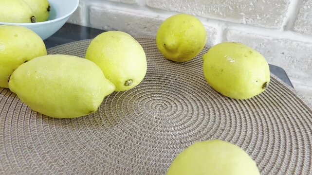 Fresh yellow lemons rolling out of a colander onto a kitchen table mat with a white brick wall in the background, showcasing vibrant, natural, and healthy citrus fruit