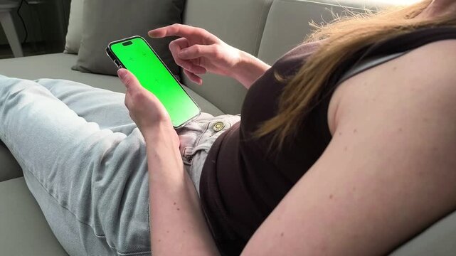Woman lies on sofa and scrolls social media on smartphone with green screen at home. Female browses feed using mobile device. Concept app promotion, digital lifestyle, online interaction,