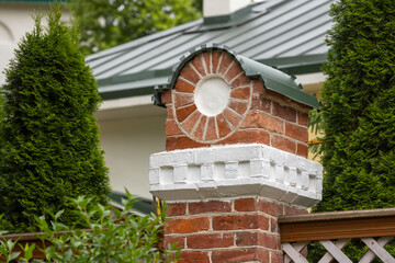 Naklejka premium Decorative red brick gatepost pillar with white plaster trim and a circular medallion, topped by a green metal cap, framed by evergreen shrubs beside a wooden garden fence of Pskov-Pechory Monastery