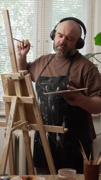 Portrait of a Caucasian, bearded millennial man wearing headphones, standing near an easel, wearing an apron, holding a palette, looking at a painting, and dancing. Concept: Hobbies and leisure