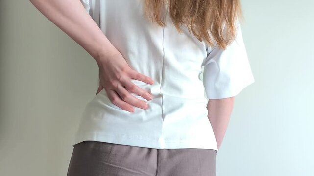 Young woman holds her lower back with one hand showing signs of pain. She presses against her spine feeling discomfort and tension in the lumbar area. Concept of backache, muscle strain and