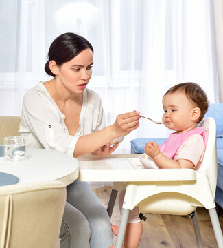 Mother feeding baby with a spoon. Spoon-feed. Happy young mother with a baby in the kitchen interior. Fresh vegetables and fruits.