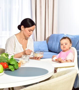 Mother feeding baby with a spoon. Spoon-feed. Happy young mother with a baby in the kitchen interior. Fresh vegetables and fruits.