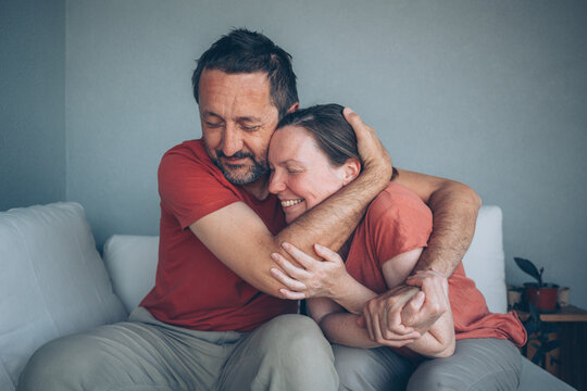 A middle-aged couple sits on a couch, embracing with warm smiles and closed eyes, expressing affection, closeness and emotional support in a relaxed, intimate home setting.