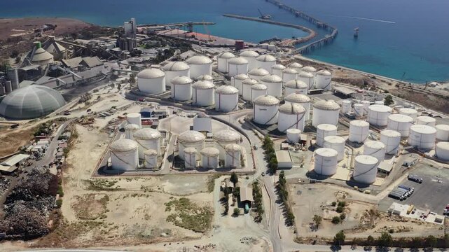 Aerial view of a large coastal industrial complex in Cyprus featuring numerous white oil storage tanks, a cement factory with silos, a long pier with port facilities, and undeveloped dry land
