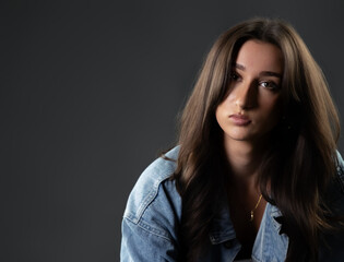 Fototapeta premium Youthful female model portrait, fashion and beauty concept, young woman with long brunette hair wearing a denim jacket. Studio shot with dark background and commercial lighting, half body view with co