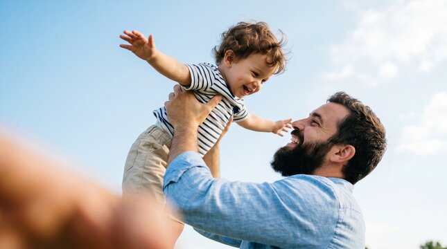 Pai barbudo sorridente levanta o filho com cabelo cacheado no ar sob o c&eacute;u azul