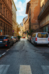Fototapeta premium Urban street scene with parked cars along a narrow road between historic buildings in Budapest Hungary during daytime with sunlight hitting architecture