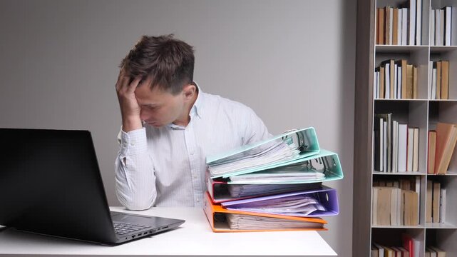 Stressed young man sitting at a desk with a laptop and a large stack of documents in binders, feeling overwhelmed and frustrated by the demanding workload and office pressure