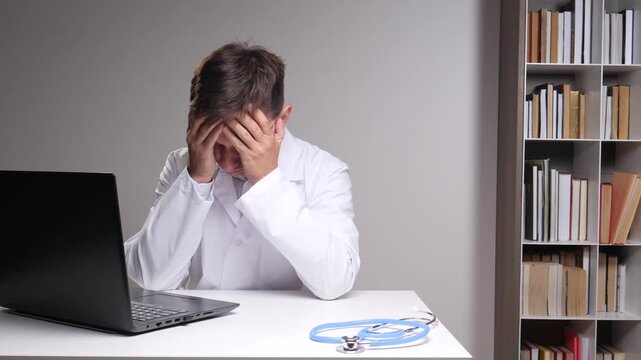 Doctor in a white coat sitting at a desk covering face with hands showing signs of stress, fatigue, and professional burnout while working from home with a laptop and stethoscope