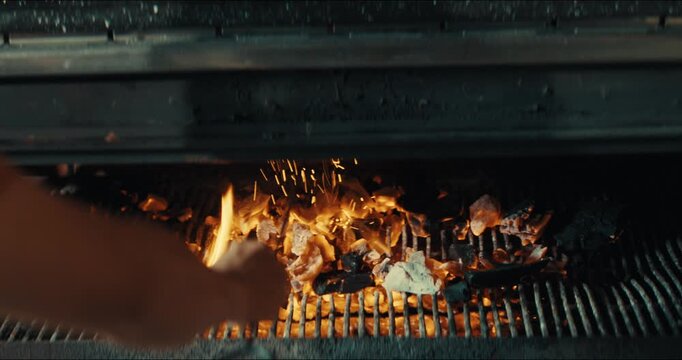 Close-up of hand adjusting charcoal on grill. Flames and sparks rise from hot coals. Embers glow beneath metal grates. Perfect for cooking demonstrations. Ideal for outdoor barbecue content
