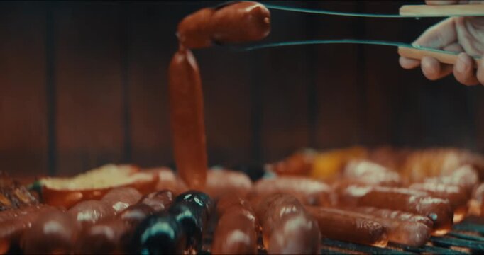 Close-up: tongs hold a glistening sausage. Sausage glows golden brown on the grill. Background shows blurred wooden planks. Hand grips tongs steadily and carefully