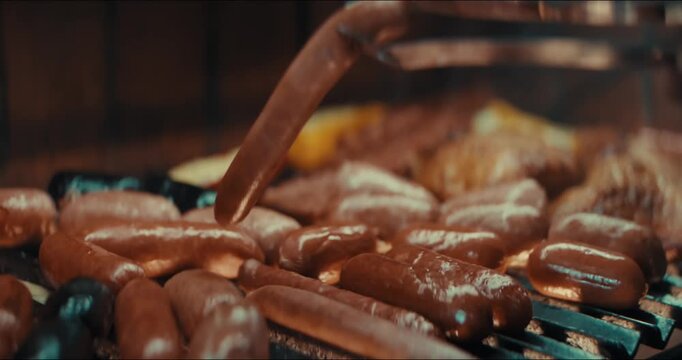 Close-up shows hand flipping hot dogs on the grill. Golden-brown sausages sizzle over the fire. Another angle features glistening meat cooking. Perfect for food videos or restaurant promotions