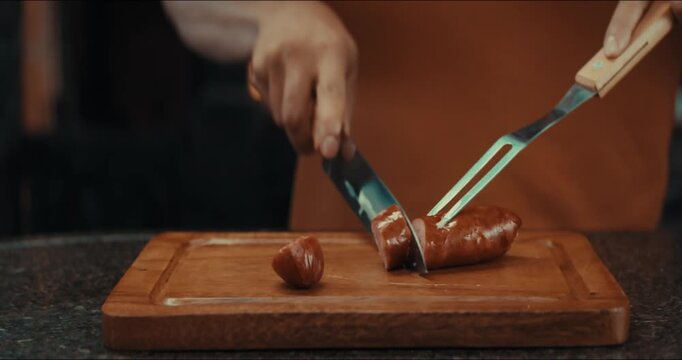 Hand slices sausage on wooden cutting board. Blue knife cuts through meat cleanly. Fork rests beside the pieces. Person's calm focus is steady. Ideal for cooking demonstrations