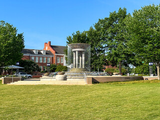 Obraz premium A modern fountain anchors a landscaped plaza at Downtown Cary Park, framed by mature trees, open green space, and historic downtown architecture.