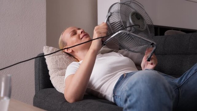 Woman suffering from extreme heat at home during abnormal heat wave, lying on sofa and using portable electric fan to cool her face in overheated indoor space.extreme heat, climate change heatwave