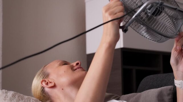 Content woman lying on sofa at home, cooling herself with small electric fan during heatwave and smiling with relief as fresh air blows on her face