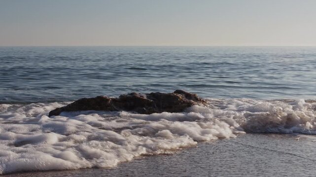 Slow motion video of crystal clear sea waves crashing and splashing against a rock on a sandy beach in Kato Gouves, Crete, Greece. Powerful nature movement