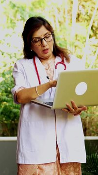 Indian woman doctor walking with laptop discussing case with young beautiful patient in hospital porch, highlighting digital consultation, patient care, wireless workflow, senior medical expertise
