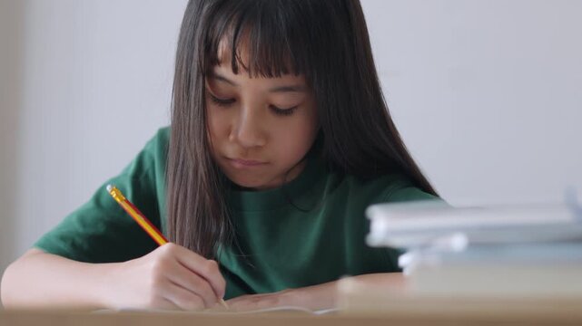 Close up selective focus of a concentrated Asian kid, a young Thai girl or Chinese student, diligently doing homework and writing with a pencil in a notebook. Education, learning, and study concept.