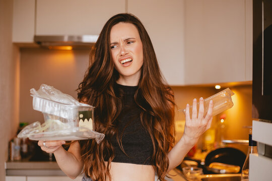 Disgusted and annoyed young woman looking away with look of disapproval, holding clear plastic containers for recycling in messy kitchen, feeling repulsed by packaging waste and domestic clutter.