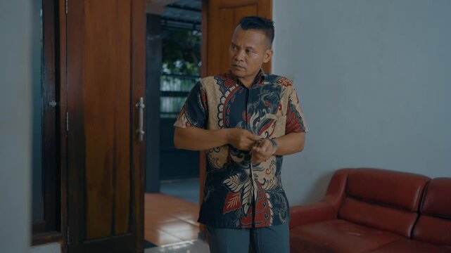 Adult Asian man buttoning batik shirt in home.