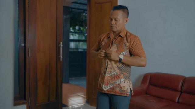 Adult Asian man buttoning batik shirt in home.