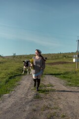 Obraz premium Young woman joyfully runs down a dirt path in a lush green landscape with a curious cow in the background during a sunny afternoon