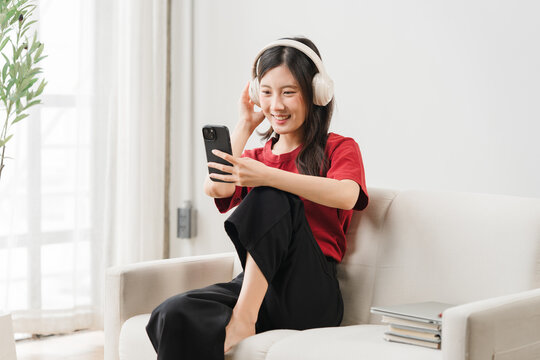 Relaxing with Music: A young Asian woman enjoys her favorite tunes while watching videos on her smartphone, comfortably seated on a soft sofa in a bright, modern living room.