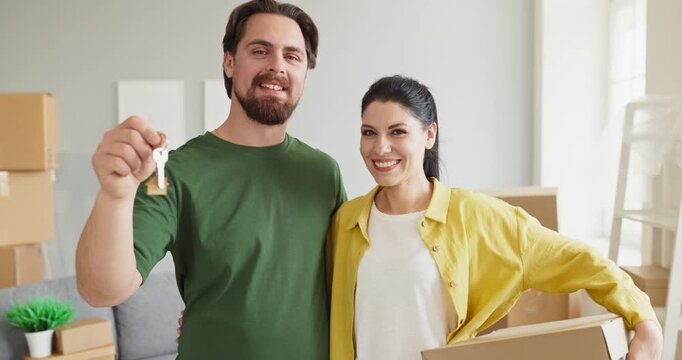 Couple homeowners hold key amid moving boxes. Smiling man and woman in a sunlit home after relocation, starting unpacking and settling in. Symbol of home purchase and new beginnings