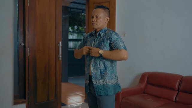 Adult Asian man buttoning batik shirt in home.