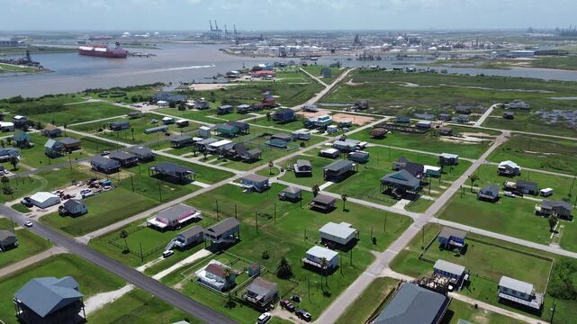 Waterfront housing reach neighborhood near Seashell Dr stretches toward inlet where cargo vessel industrial facilities sit along distant shoreline. Clean roof patterns, open grass, Freeport, TX