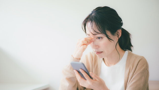 Stressed Japanese woman looking at smartphone with tired eyes スマホを見て目が疲れ頭を悩ませる日本の女性