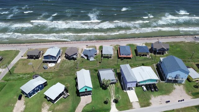 Beachfront stilt row homes near Seashell Dr line Gulf corridor beside narrow road rocky barrier separating shoreline from residential edge. Varied roof colors, grassy setbacks, surf, Freeport, TX