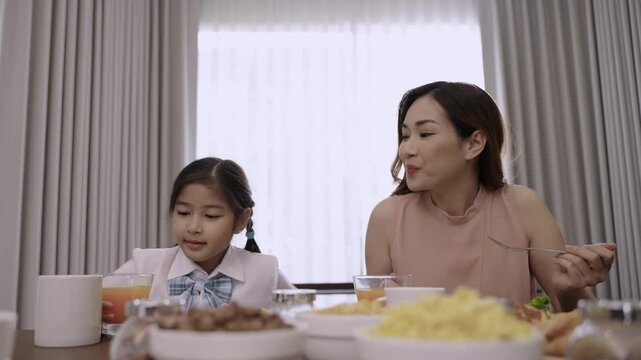 Happy Asian family mother and child daughter in school uniform sitting smiling while eating breakfast and drinking orange juice for health together before school during morning in the kitchen at home.