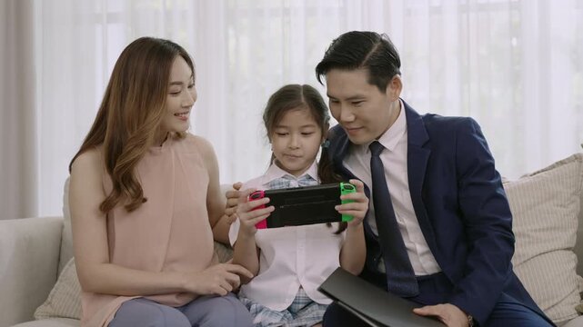 Happy Asian family, Mother, and Father sitting relaxing and looking at their daughter playing portable game console with joyful expression on sofa in living room at home. Dad in suit return from work.