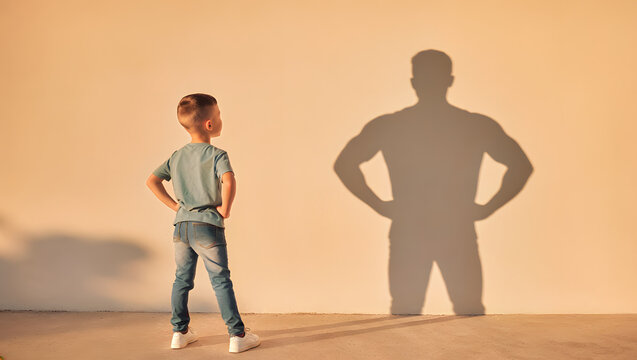 A little boy standing with hands on hips looking at his shadow cast as a strong muscular adult man on the wall, symbolizing future potential, growing up, ambition, and confidence.