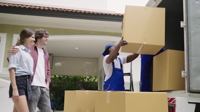 Happy young Caucasian couple relocating, standing, looking, and checking with professional movers in uniform loading items and furniture onto a truck for moving and delivery to the new home. 