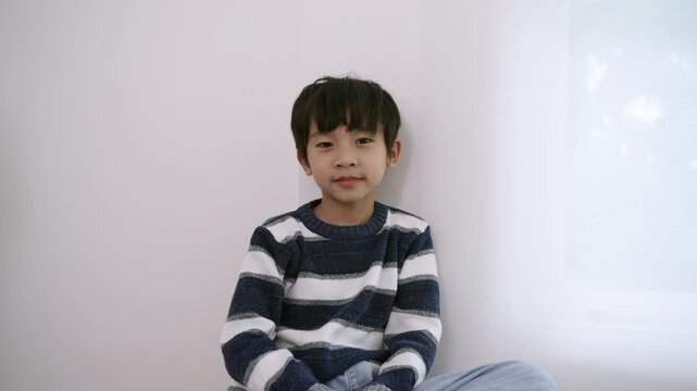 Happy, adorable Asian boy sitting smiling, raising his hand, saying sawasdee, and paying respects in the Thai style, waving to the video camera while playing and looking at the camera in homeschool. 