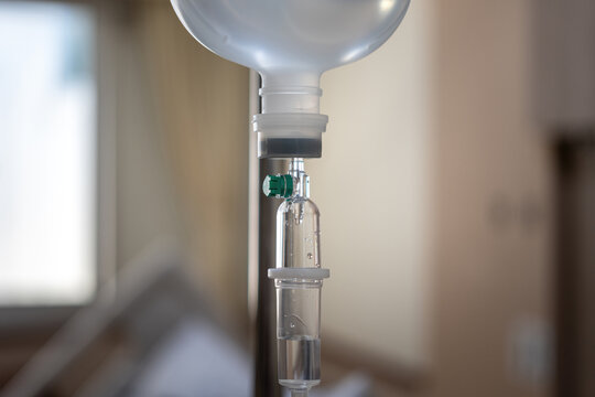 	
A close-up shot of an intravenous (IV) drip infusion set with saline solution drops in a hospital room, featuring a blurred background.
