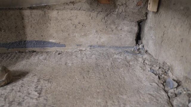 Worker pours yellow liquid onto concrete floor near wall crack. Liquid spreads across rough surface. Construction repair or waterproofing process shown