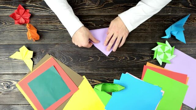 Top view of a person folding a purple paper sheet on a dark wooden table with origami figures