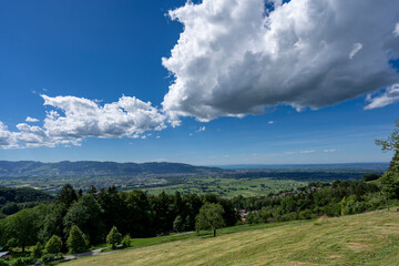 Fototapeta premium Panoramic high-angle view over the lush green Rhine Valley in Vorarlberg, distant mountains of Switzerland, and Lake Constance under a vibrant blue sky with dramatic cumulus clouds