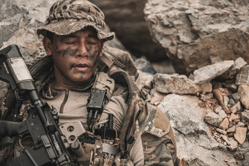 Obraz premium Exhausted military commando resting on rocky terrain during a combat mission. Tired armed forces operator catching his breath after battle. Tactical survival and warfare concept.