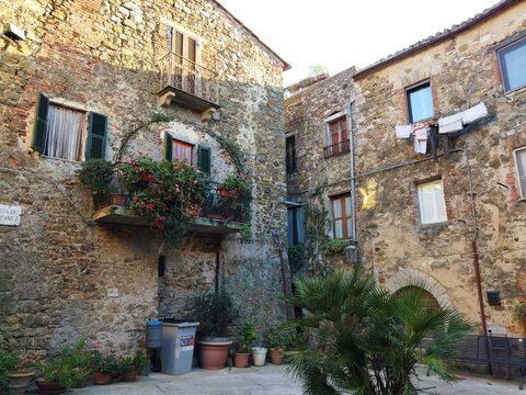 Plaza medieval con fachadas de piedra hist&oacute;ricas, balc&oacute;n decorado con flores y ropa tendida en Montemerano, Toscana, Italia.