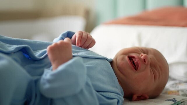 Unhappy newborn baby boy in blue pajamas lying on his back on a bed, crying and expressing discomfort. Adorable and vulnerable infant making grimaces and moving his tiny hands in distress
