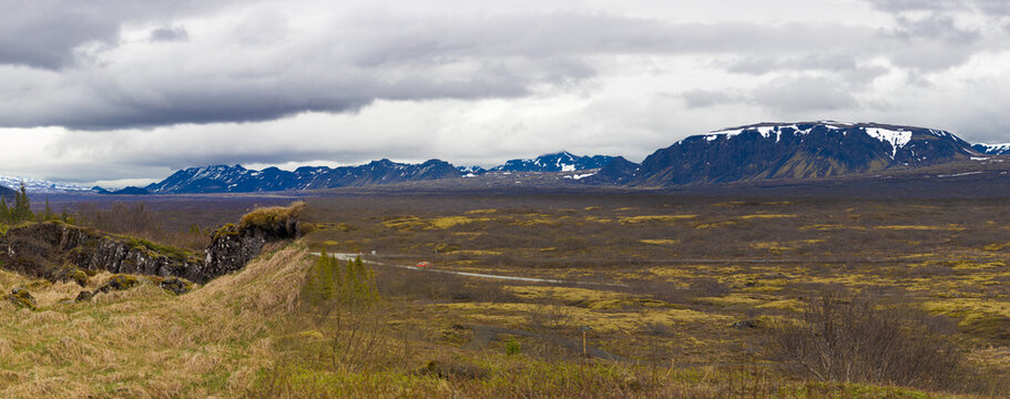 Wide panoramic view of Thingvellir National Park in Iceland under a cloudy sky, showcasing vast mossy tundra plains and an orange vehicle on a distant road against snow-capped mountains.