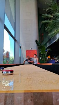 A low-angle shot of a table shuffleboard game with shiny pucks on a long wooden surface, featuring two people playing in a modern indoor space