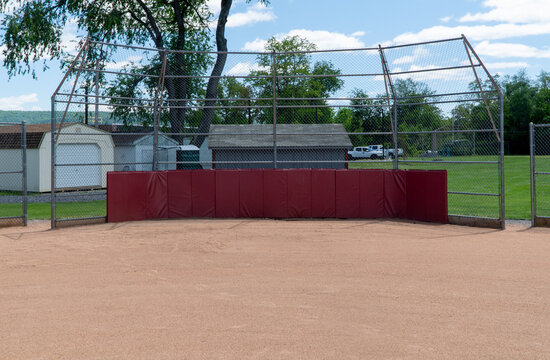 Empty baseball or softball field with a protective chain-link backstop and red padding, featuring a dirt infield, green outfield, sheds, and blue sky with clouds on a sunny day.